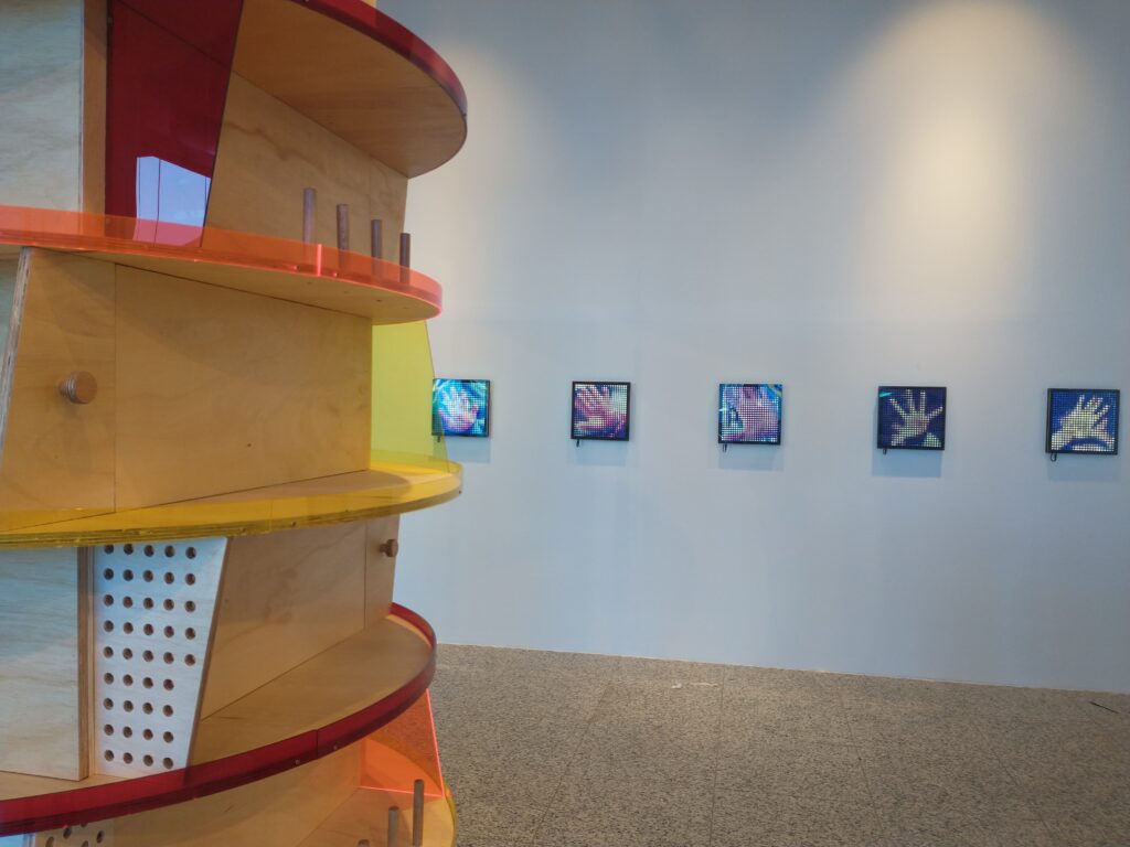 Imagem de parte da exposição Na Palma da Mão com uma bancada circular em madeira e acrílicos coloridos, tendo ao fundo quadros.
