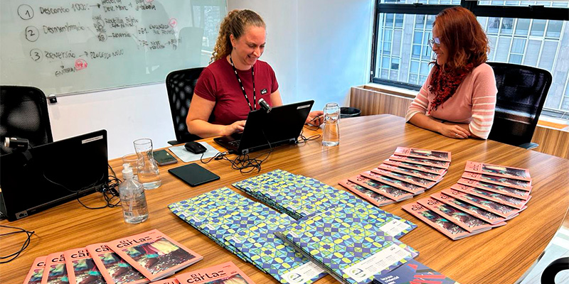 A imagem mostra uma funcionária do Sesc sentada em uma sala de reuniões, interagindo em frente a outra mulher, enquanto tecla um laptop. A mesa de madeira está organizada com materiais impressos, Revistas E e Guias Em Cartaz. No fundo, há um quadro branco com anotações e uma janela grande que deixa entrar luz natural, revelando uma vista urbana. Além disso, há itens como garrafas de água, copos e álcool em gel sobre a mesa.