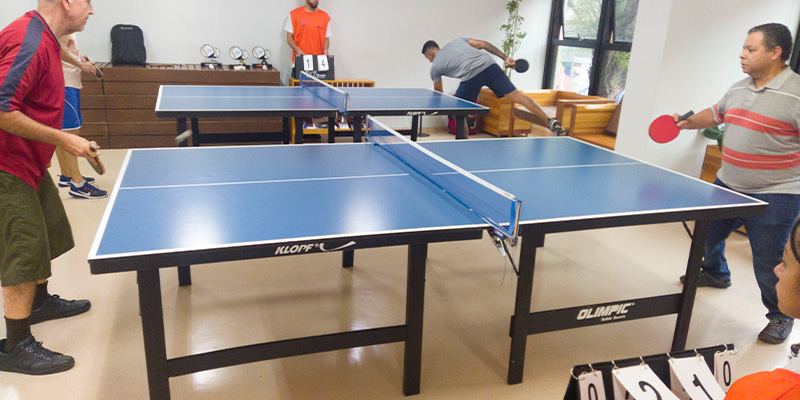 Duas mesas de tênis de mesa montadas na Área de Convivência do Sesc Carmo, um espaço coberto com poltronas de madeira ao redor. Pessoas jogam competindo em torneio da Copa Sesc.