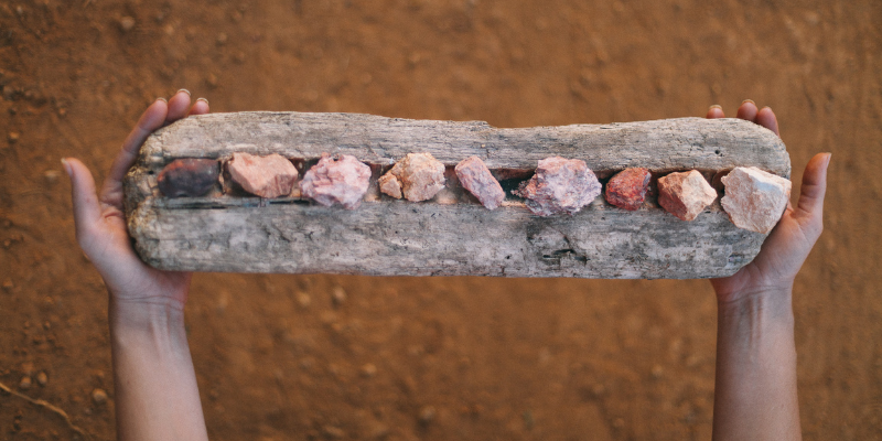 Fotografia mostra mãos de pessoa branca segurando um objeto artístico, que consiste em uma espécie de prancha de madeira sobra a qual estão nove pedras coloridas.