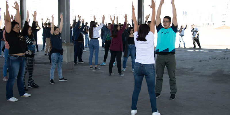 A imagem mostra um grupo de pessoas participando de uma atividade física coletiva em um espaço coberto e amplo. Os participantes, distribuídos em várias fileiras, estão com os braços levantados, seguindo as orientações de um instrutor que está de frente para eles. O instrutor veste uma camisa azul e calça verde, enquanto os demais usam roupas casuais. A atividade parece promover alongamento ou exercícios leves, como parte de um evento de bem-estar ou integração.