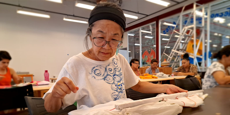 descrição resumida: Miriam Fukai , mulher amarela, de cabelos grisalhos, presos, usa uma faixa de cabelo preta, óculos de aro quadrado transparente, e camiseta branca com desenhos de arabescos em azul. Ela está diante de uma mesa cinza, com rolos de linha e borda em um voil de 1metro. Ao fundo a sala do ETA casa verde. 
